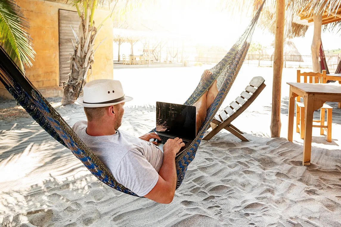 A man in a white t-shirt sitting on a hammock. He is typing on a laptop. In the background, a beach and wooden furniture are visible.