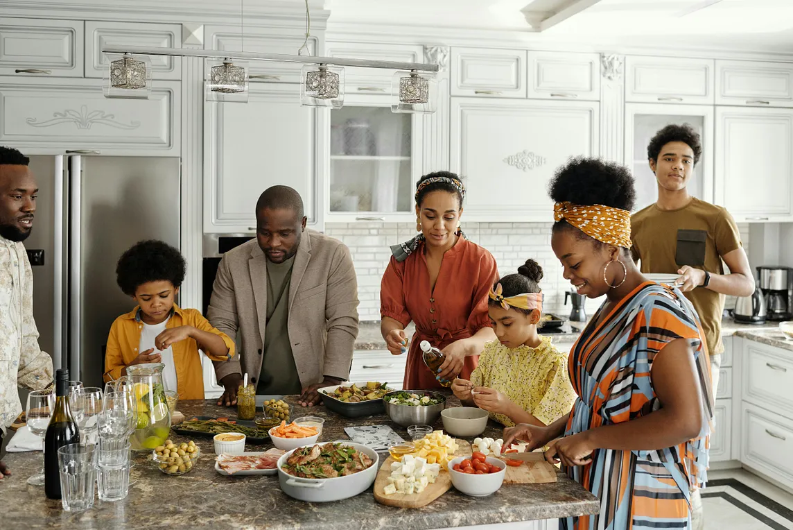 An extended family of Black people in a well-appointed kitchen preparing food.