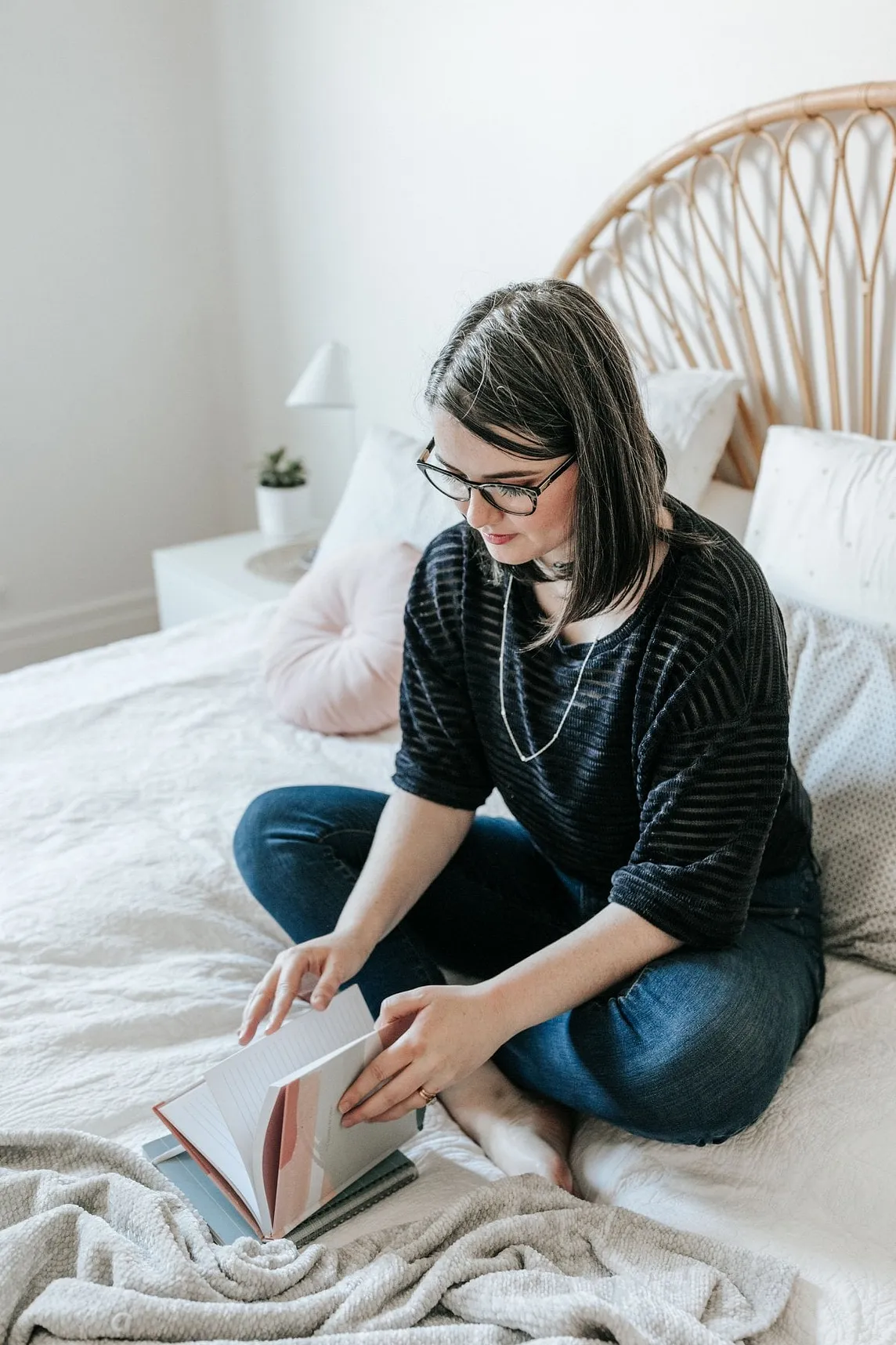 Lara, a white woman with glasses and a mid length bob of dark hair, holds a book with lined paper while sitting on a white bed. She is looking down at the book and wearing a blue striped blouse and jeans.