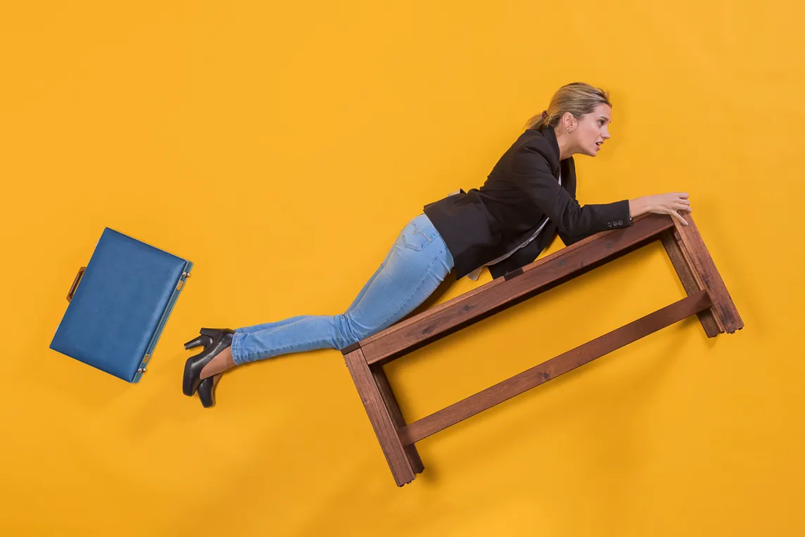 A person in business attire (black blazer and jeans) lies stretched across a floating wooden bench against a bright yellow background, reaching forward while a blue briefcase appears to fall away in the opposite direction. The surreal, gravity-defying composition suggests career transition or letting go of traditional corporate path