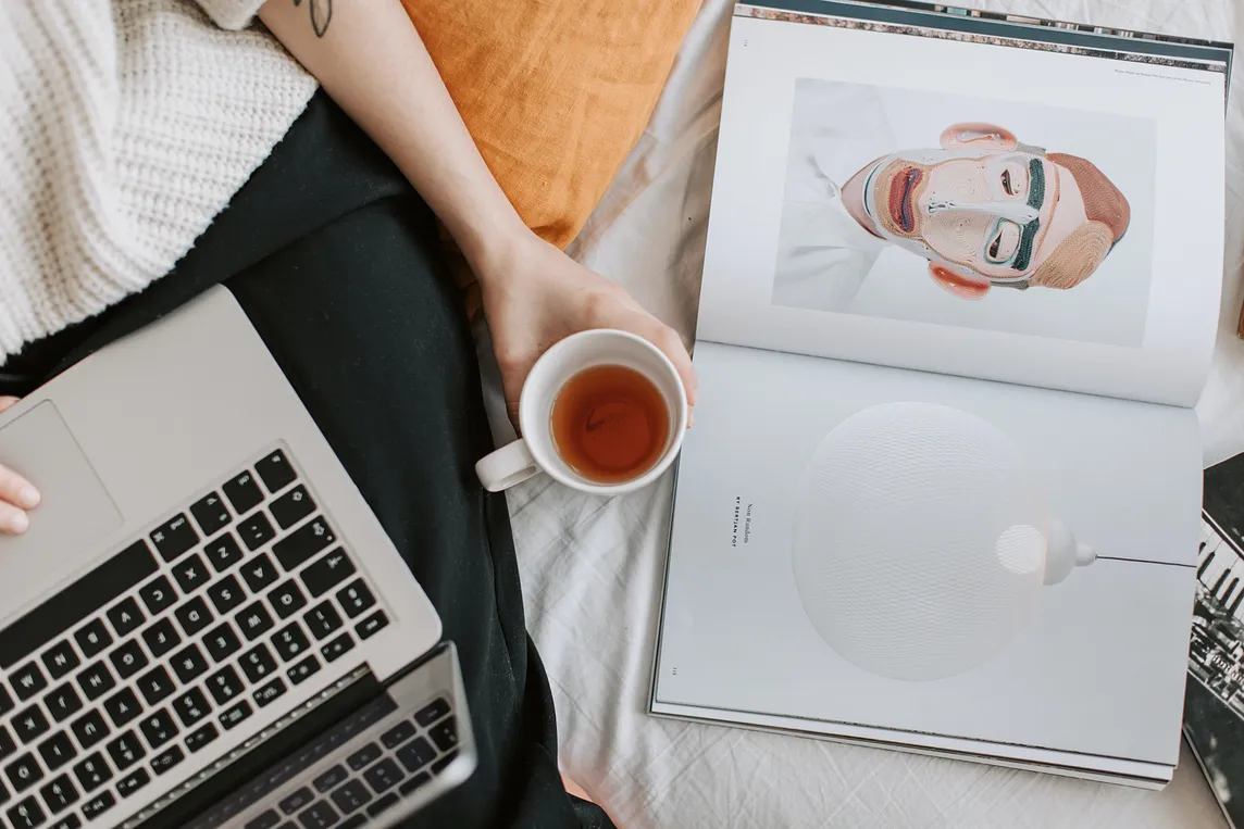 A woman siping tea reading a book on her macbook.