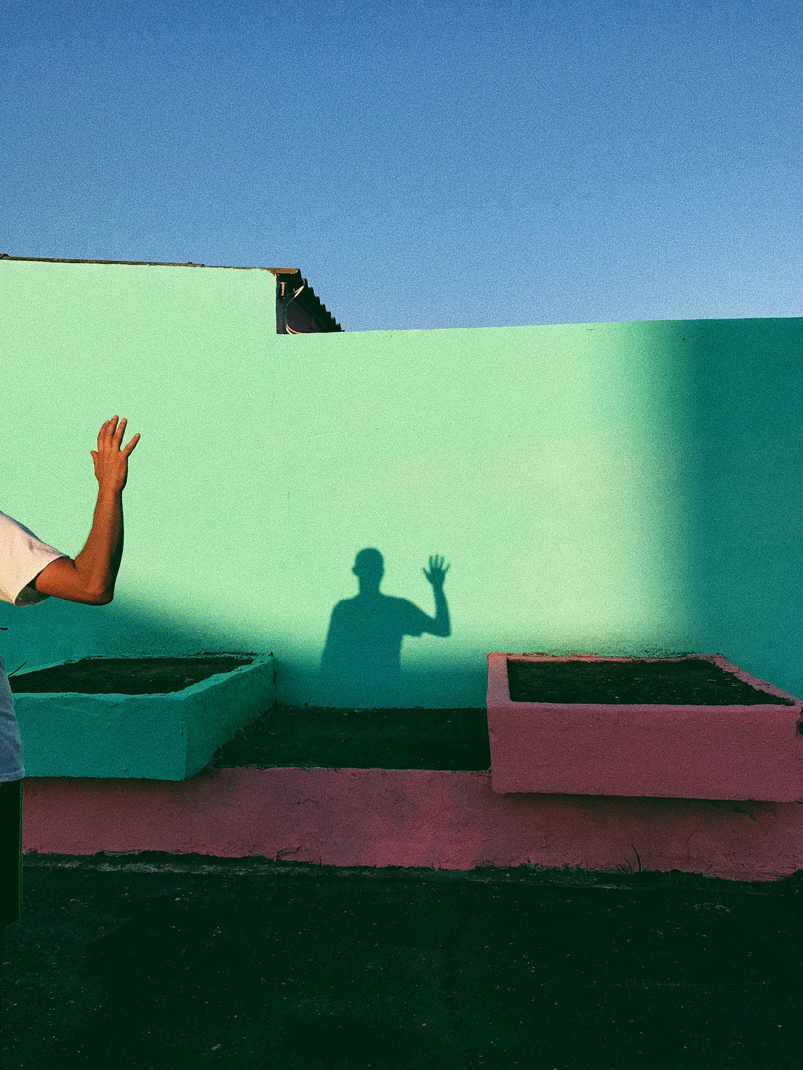 Person in foreground with only one raised arm visible, in the background the shadow of the person’s raised arm and torso are seen on a teal wall.