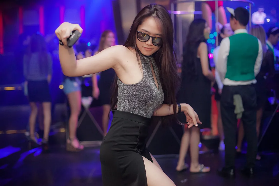 Image of young woman, long black hair, sunglasses and sparkly top dancing in a nightclub, holding phone to take a picture. Background shows other people in club