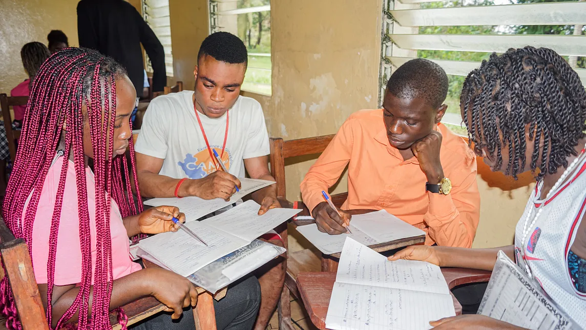 Four students sit facing each other at school desks covered with notebooks they are writing in and studying.