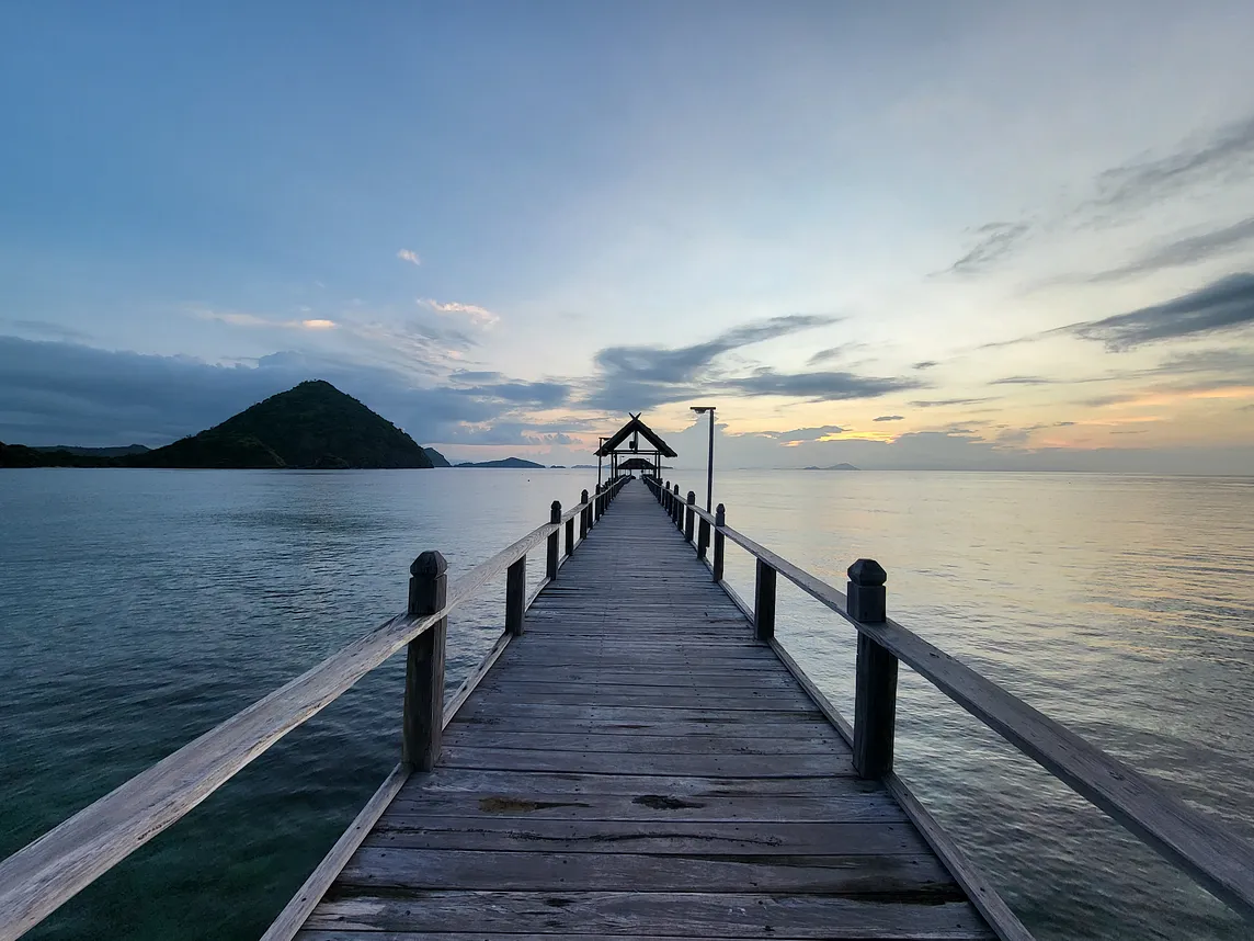 A wooden pier on the sea, leading to a sheltered gazebo, set against a backdrop of a sunset and mountain.