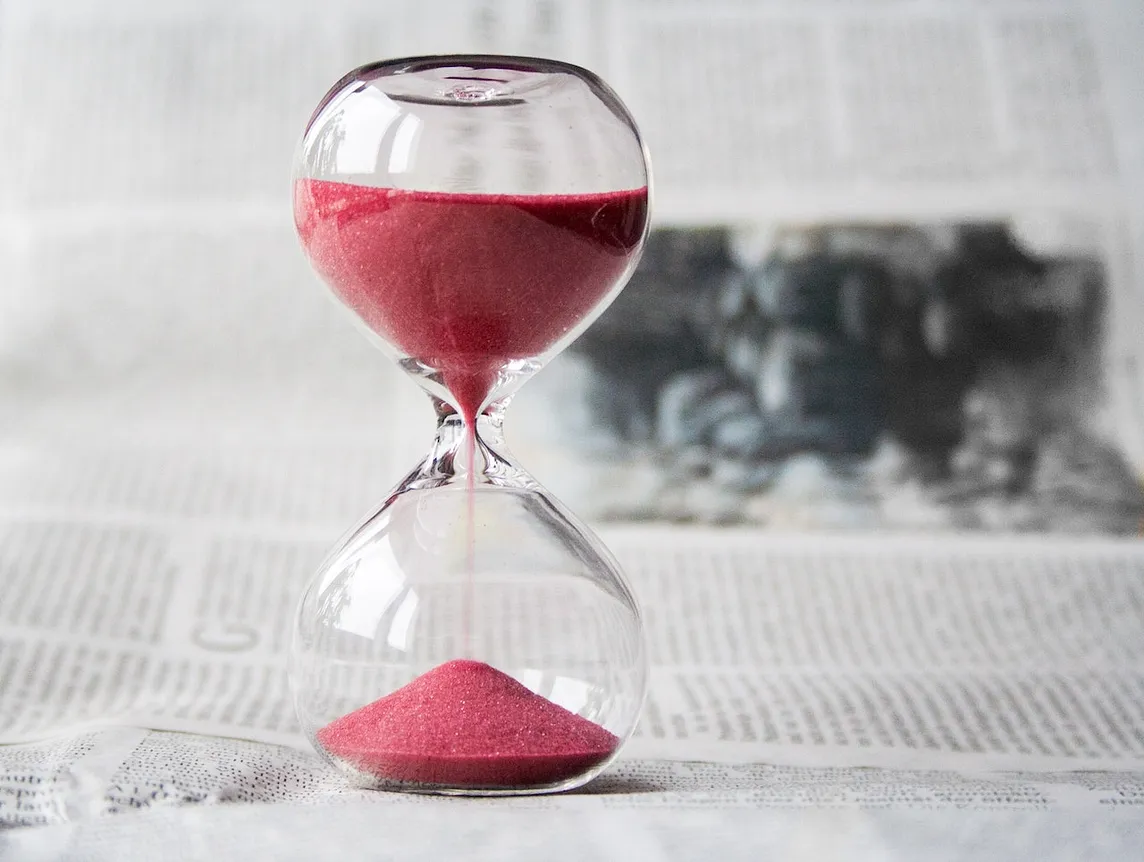 Clear glass hourglass with red sand running through it against a greyed out backdrop of printed paper.