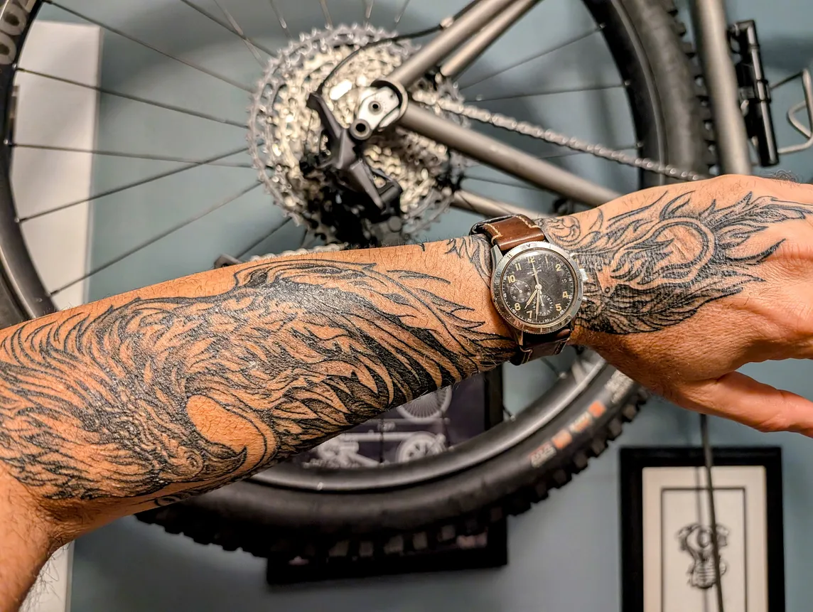 Closeup of the author’s forearm and hand with a black and grey tattoo of a firebird. He’s wearing a windup watch with a leather band and there’a bicycle wheel in the background.