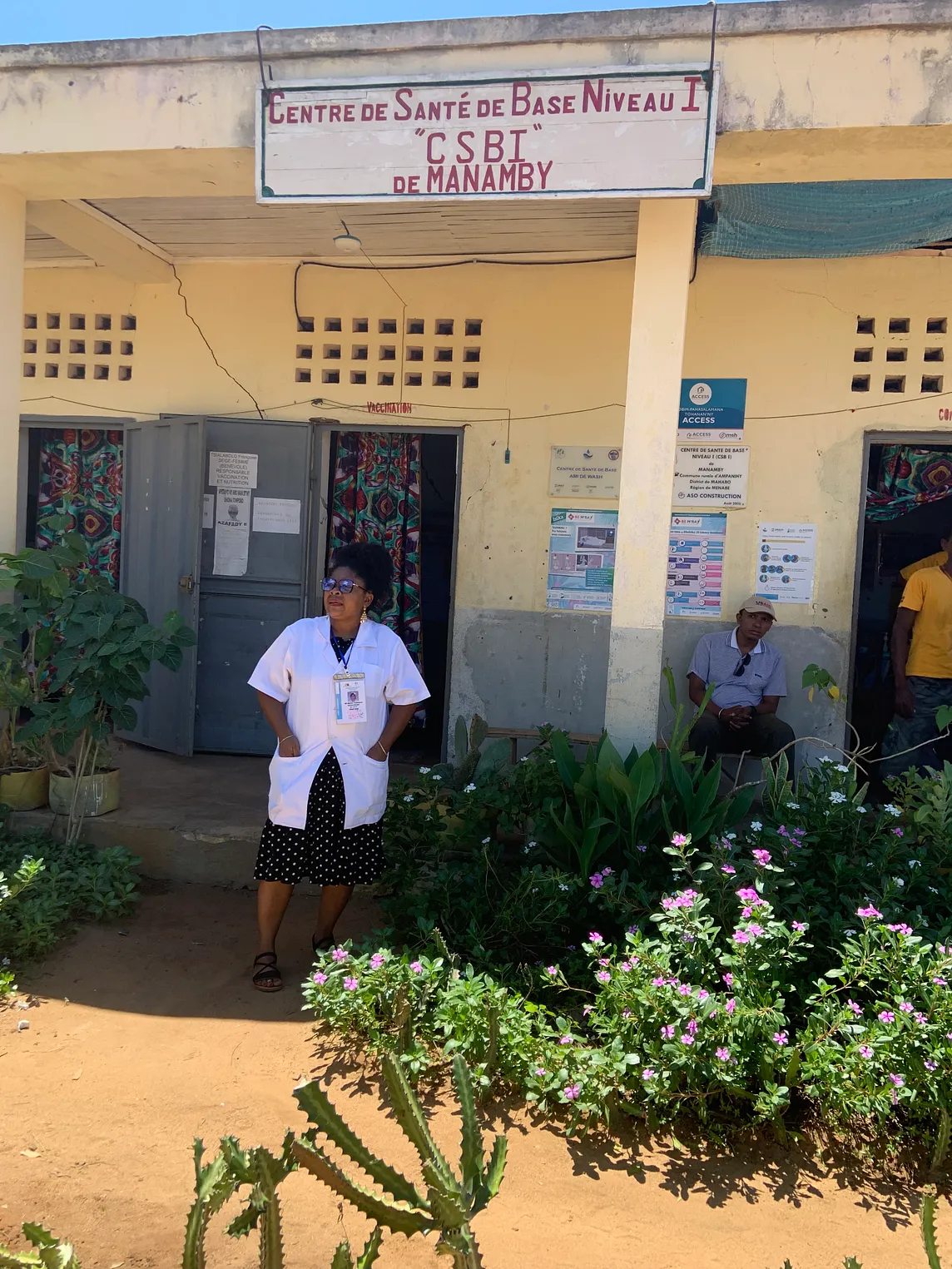 A nurse-midwife wearing a white lab coat stands in front of a community health center with an entrance adorned with lush flowers and greenery.