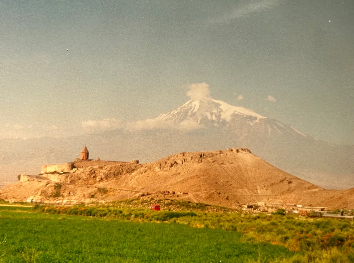 Large snow-capped mountain rises in the background. In front of it is a large hill of dirt with a small-peaked building on the left side. Fields of grass are in front of the hill, in the foreground.