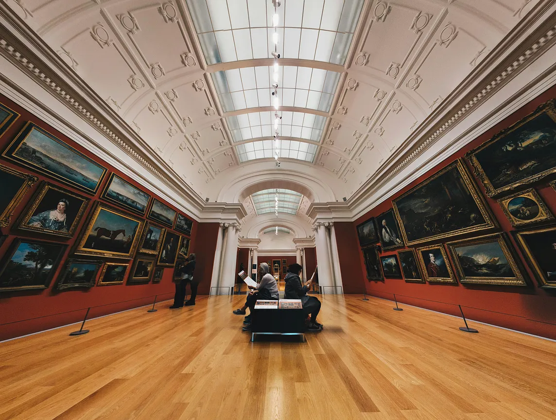 An art gallery room with high curved ceilings, parquet floor and old paintings in golden frames with a bench at the center to visitors to rest