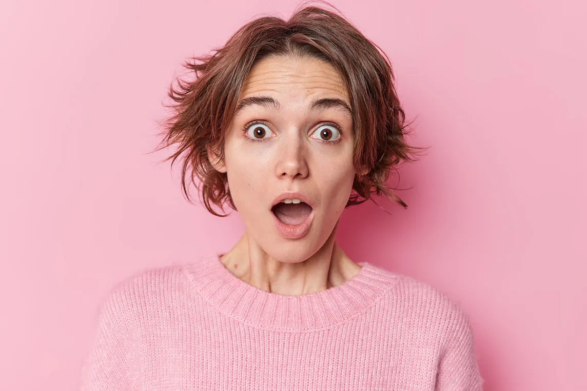 Portrait of a stupefied young woman staring into the camera with her mouth open. She looks panic-stricken. Her brown hair is a mess. She is wearing a casual pink jumper. All against a pink background.