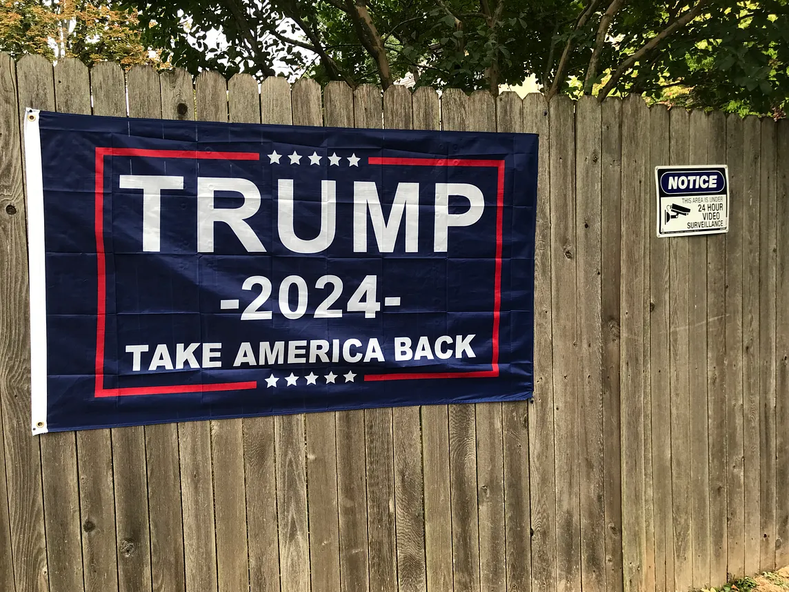 Trump sign posted on a fence beside a sign stating that the area is under video surveillance.