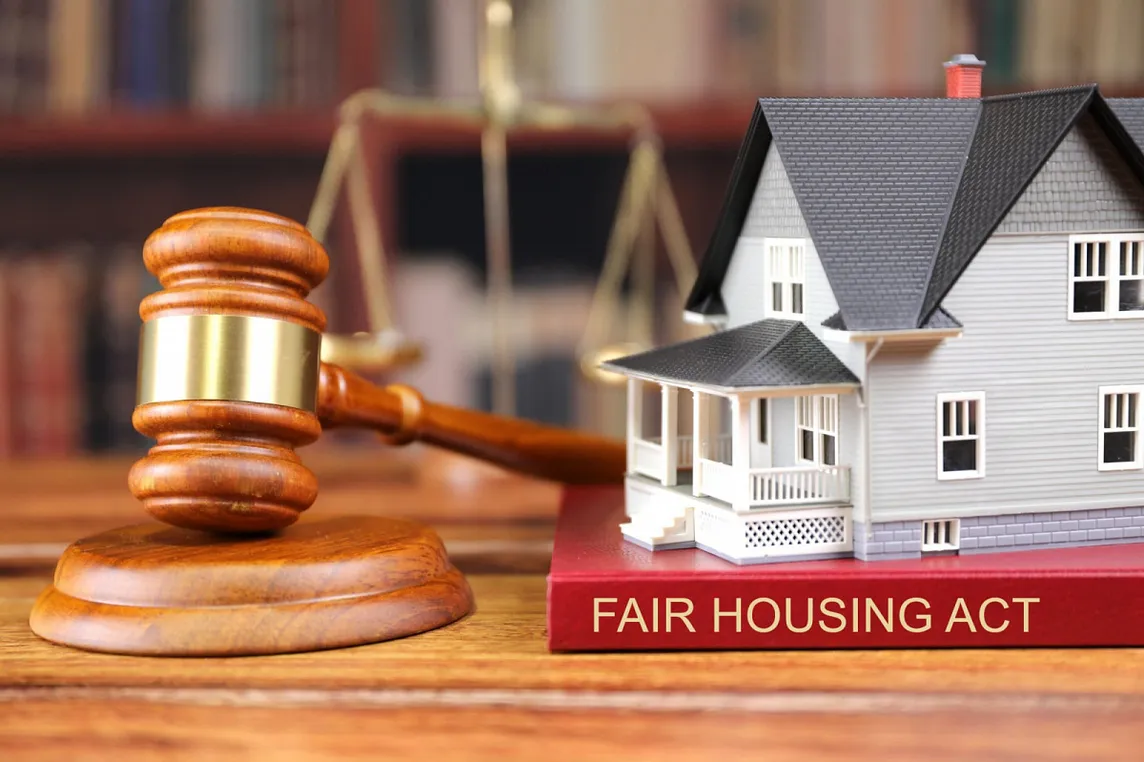A photo shows a wooden gavel propped on a wooden sound block. To the right of the gavel is a miniature model of a white and gray two story house with a covered porch in front. The  model rests on a red book with the title “Fair Housing Act” on the spine. An old fashioned scale and books on book shelves can be seen in the background, but they are blurred.