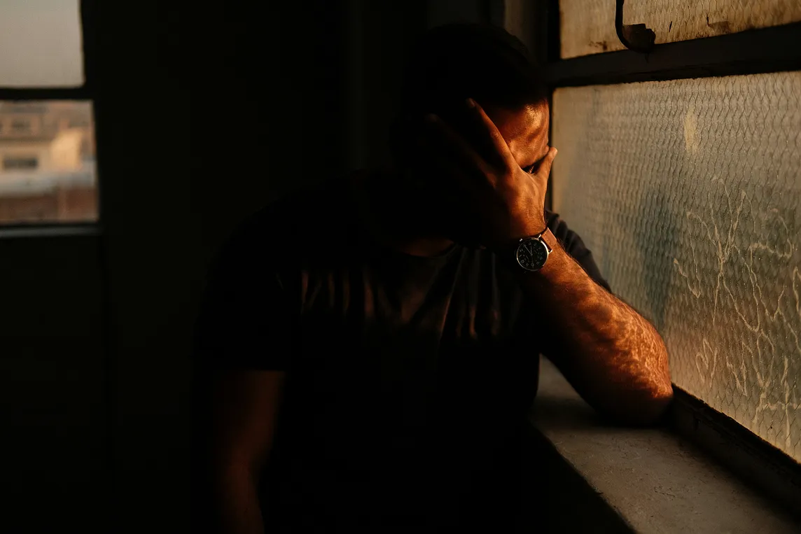 A sad man in the dark covering his face by the window