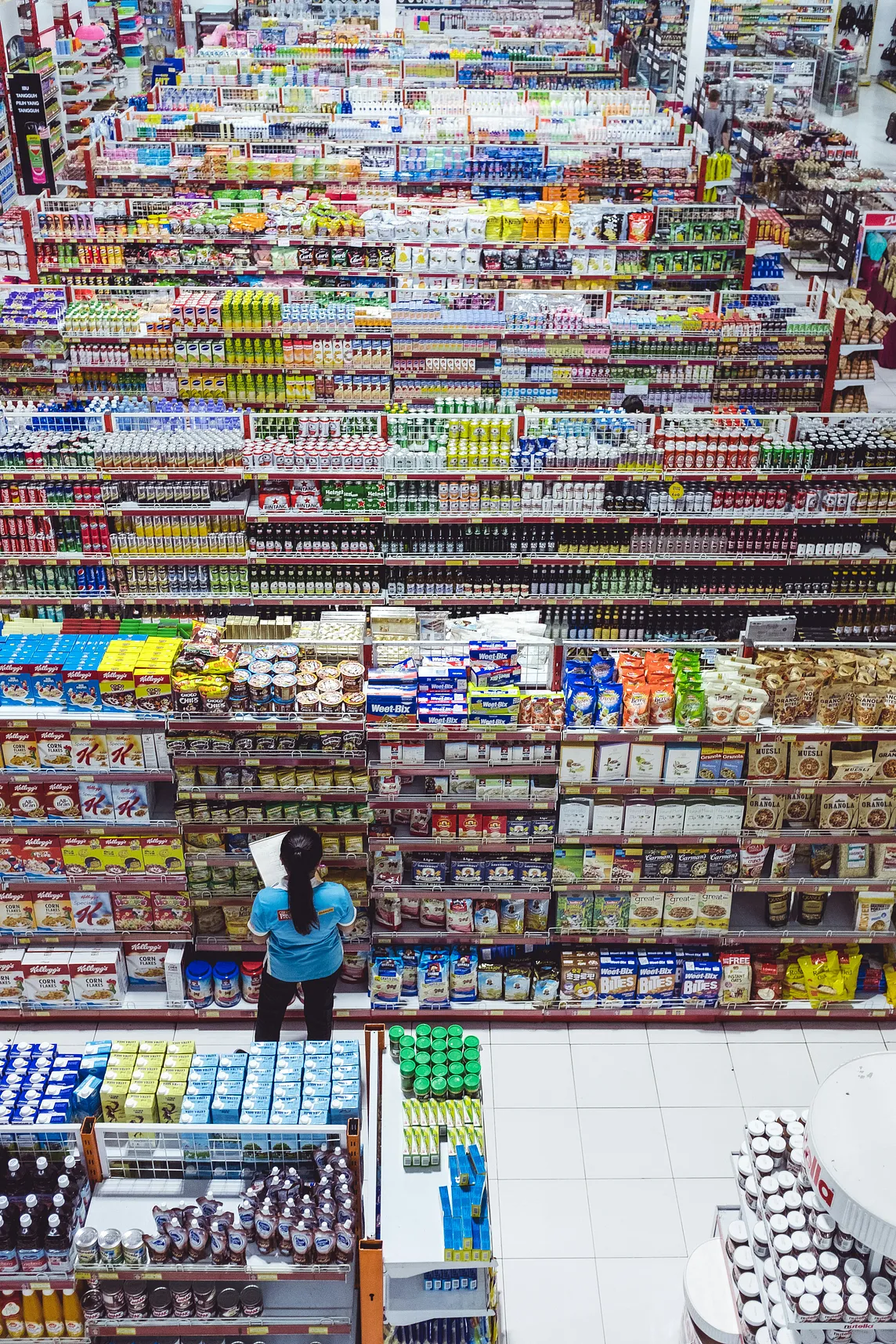 Aisles and aisles in a grocery store with woman in blue shirt and long dark hair in a pony tail standing in front.