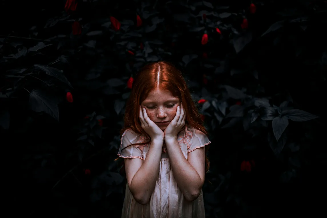 A little girl with long red hair, looking down sadly with her hands on her cheeks, against a dark background. PJ Jackelman.