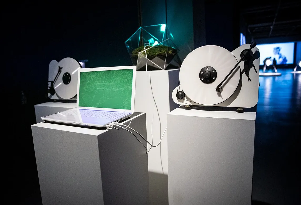 A laptop, two vinyl players and the moss installation each of them placed on a white pillar in an exhibition space.