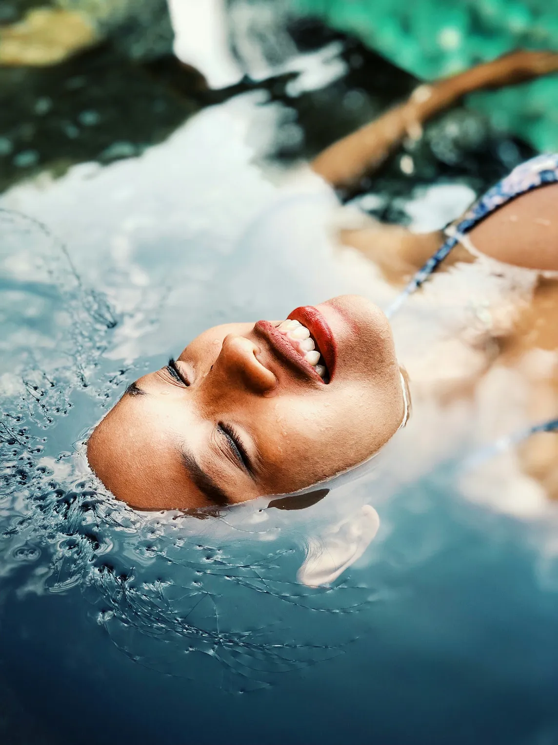 “Embodiment and Reconnecting to Self: Embrace Inner Harmony” This photo shows the face of a woman with carmel-brown skin floating in a pool of blue water. Only her face is above the water and she is smiling.