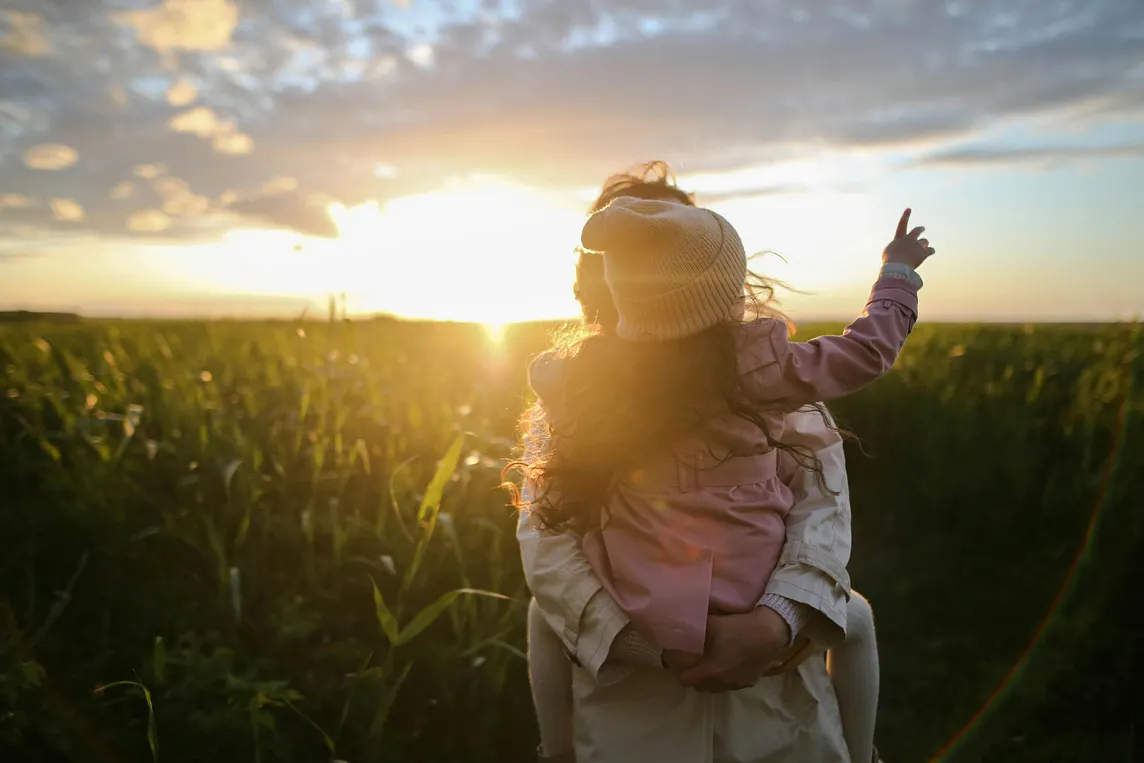 While he sun sets in the background on a field of corn, a mother holds her young child. The child is wearing a hat and pink clothing, with a hand raised in the air.