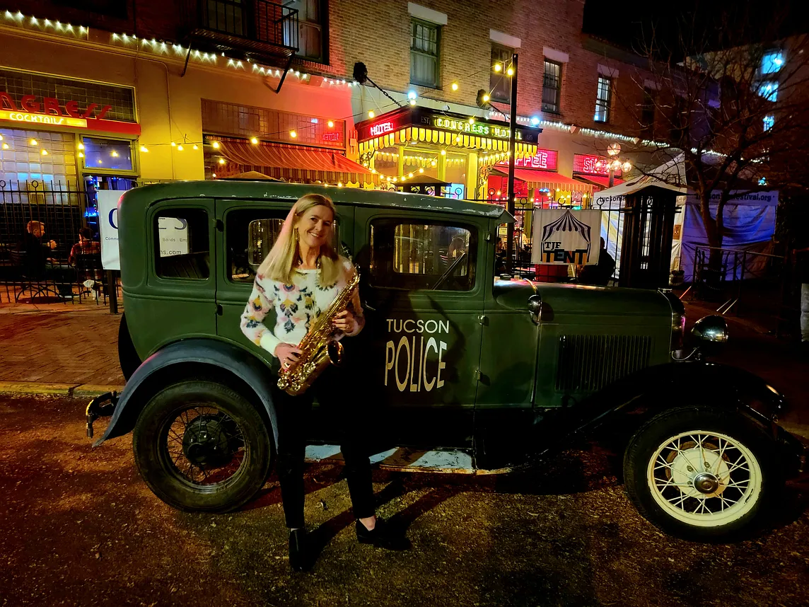 The author at night with her alto sax standing by a green Model T (?) with “Tucson Police” painted on the door. The historic Hotel Congress is in the background.