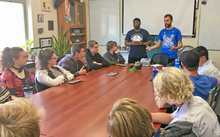 A group of students sitting at a round table with two adults standing at the head of the table.