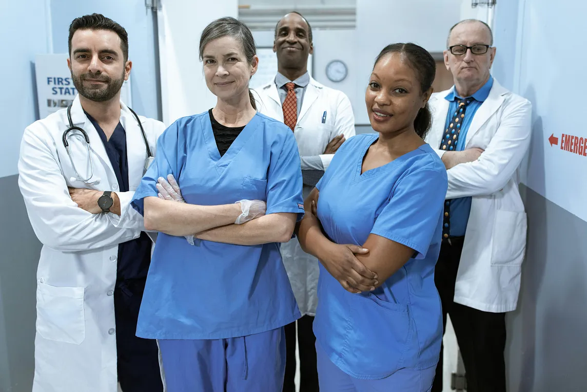 A group of medical professionals with their arms crossed in a hospital hallway. There are two women in front and three men in the back.