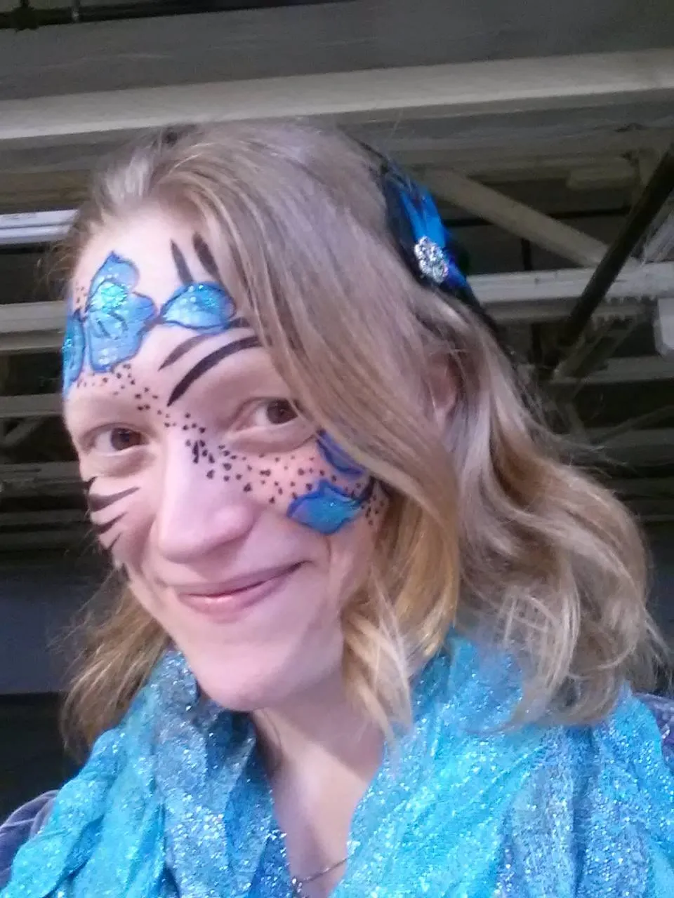 White woman with wavy strawberry blonde hair, smiling, with face paint of blue shimmery flowers, strong black lines, and a smattering of dots from forehead, swooping across the nose to the cheek. A small blue feather and silver clip is in her hair and she has a matching blue scarf on.