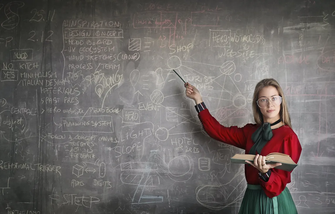 Female teacher holding an open book and pointing to complex diagrams on a chalkboard