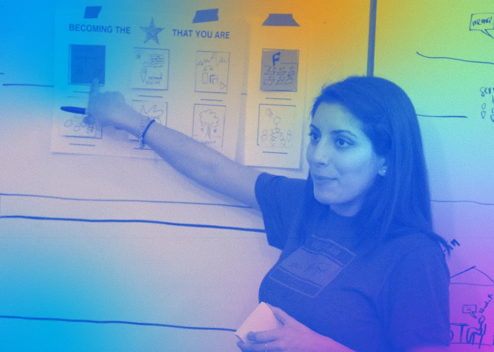 Tania Anaissie stands near a white board and points to a handout taped to its surface. The title of the handout says, “Becoming the Star That You Are.” This image is colorized with shades of light blue, gold, green and pink.