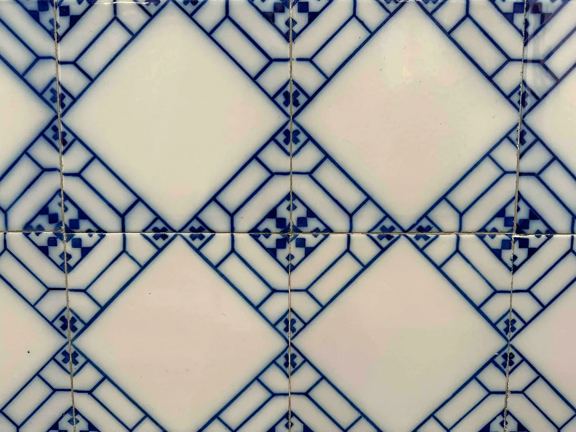 A close-up image of a tiled surface with a repeating pattern. The tiles are square-shaped with white centers and edges decorated in blue geometric and floral designs. The pattern creates a visual effect where blue diamond shapes appear at the intersections of the white squares. There are visible grout lines between the tiles, and slight imperfections can be seen on the surface, indicating wear or age.