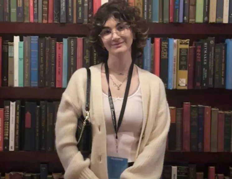 A young person wearing a pale sweater with a white shirt, glasses, black purse on their left shoulder, and a blue pass around their neck is standing in the middle of a book shelf. They have pale brown hair with some blonde streaks in the front and have a smile on.
