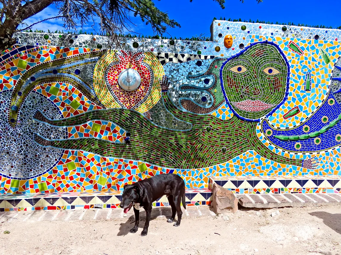 A black labrador dog stands, panting, next to a wall of mosaics illustrating a green figure swimming in a sea of color