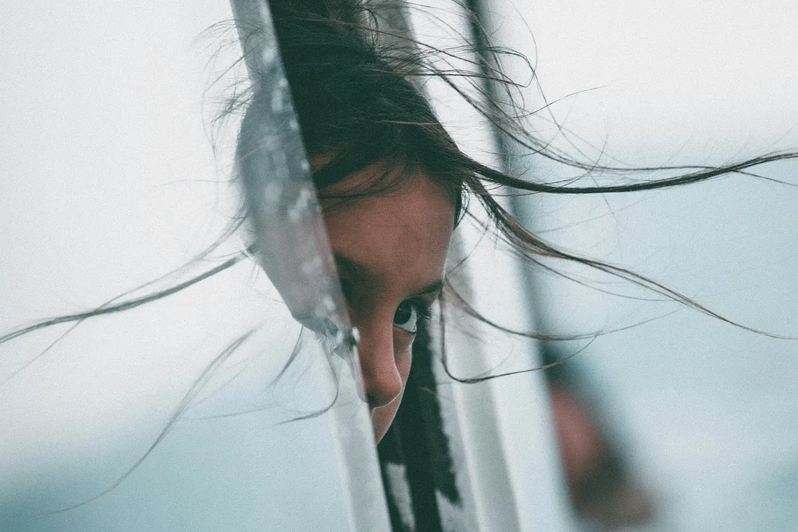 A young girl’s hair is swept up in the wind, reflected on the glass she peers behind.