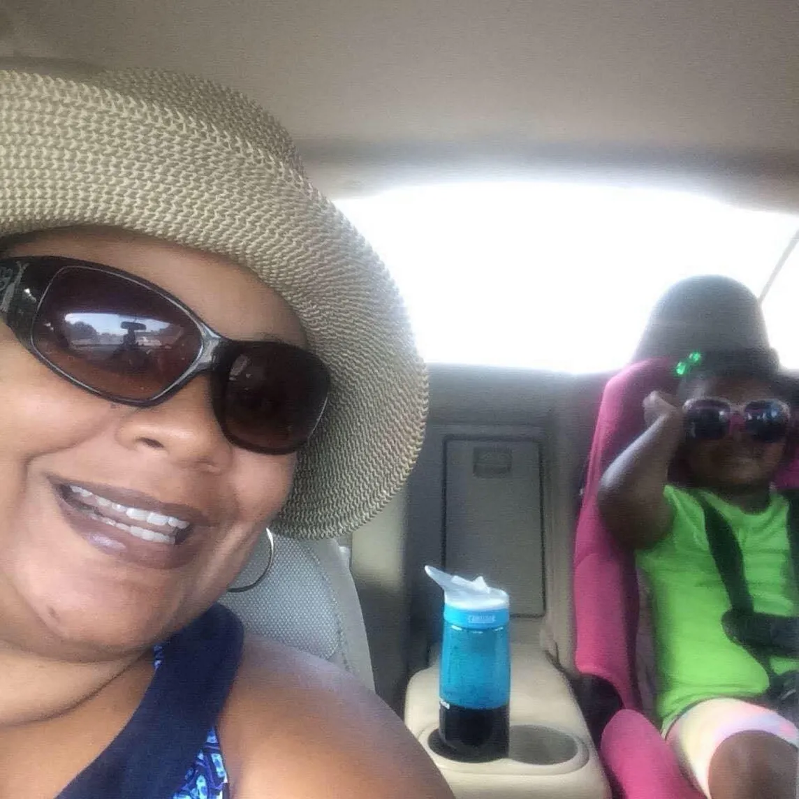 A lady wearing a hat and sunglasses in the front seat and her niece wearing shades and a green top in the back seat. Both are smiling.