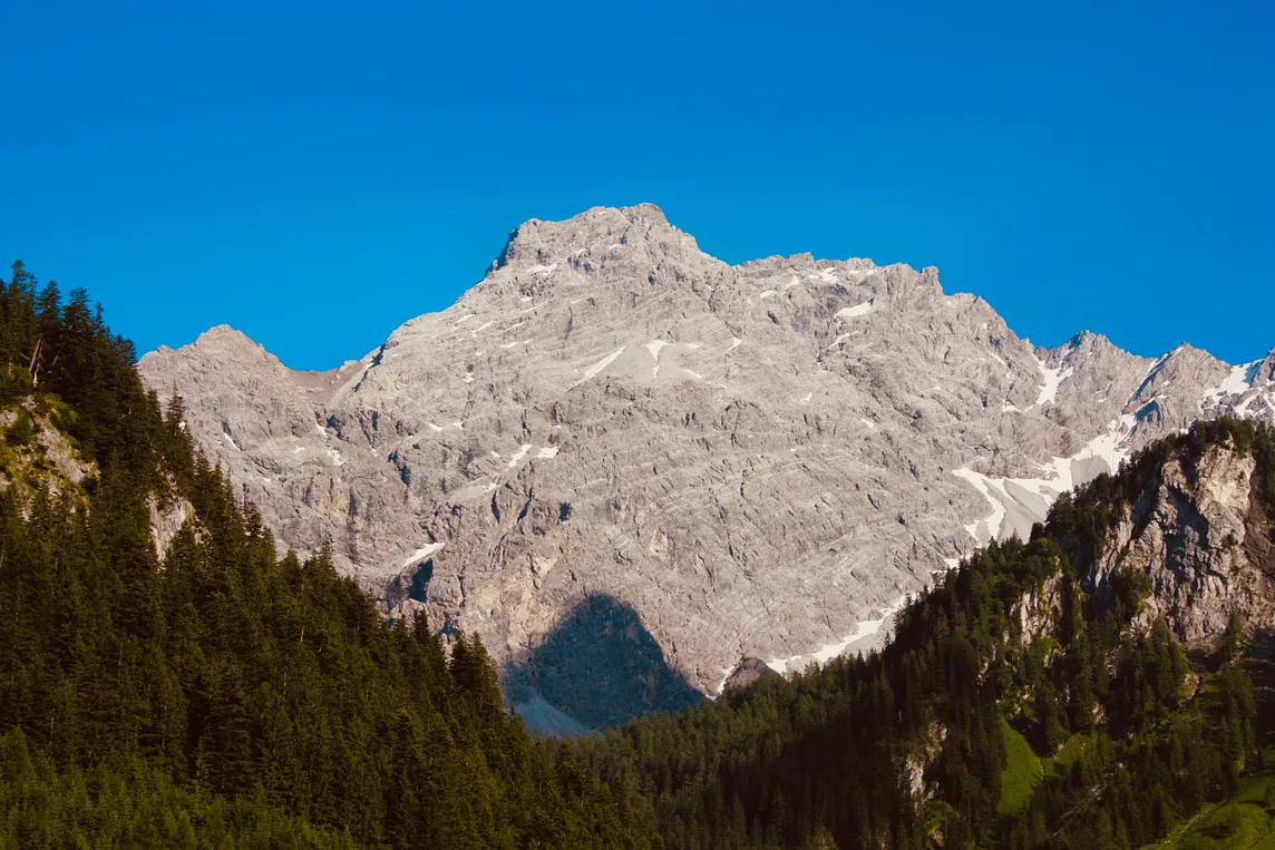 A beautiful bare-rock mountain with a few thin strips and pockets of remaining snow (the picture was taken in the height of summer) towering impressively into a deep blue sky. In the foreground the picture is framed by two steep slopes, one going left and the other right, covered in conifer forests.