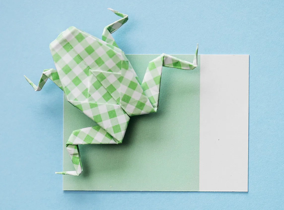 A paper origami frog made of white and green checked back sitting on a piece of green paper with a white strip on one side against a light blue background.