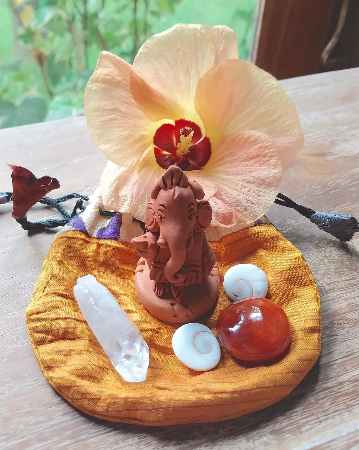 A travel altar with a clay figure of Ganesh, crystals and a big peach coloured tropical flower on a desk looking out onto a garden