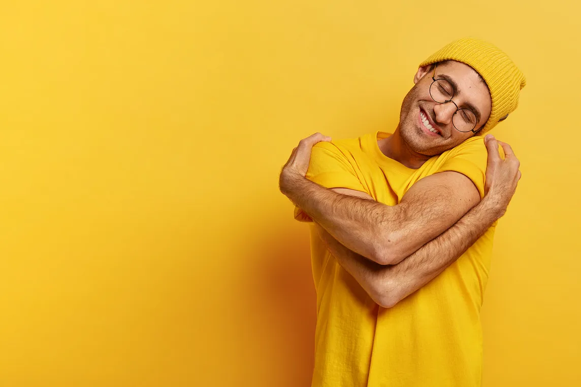 A happy man hugs himself. His head is tilted, and he has a toothy smile. He is wearing a yellow T-shirt and yellow hat against a yellow background.