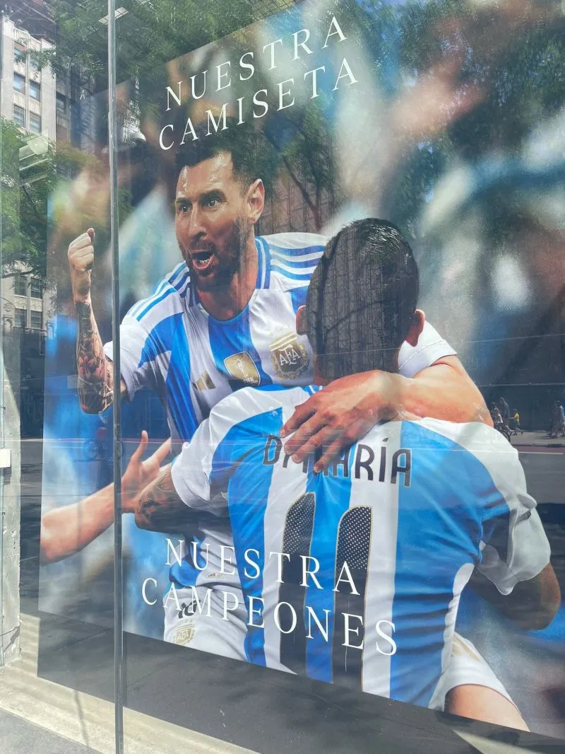 Poster of Messi giving teammate celebratory hug on soccer field, text on the advertisement reads “Nuestras Camisetas Nuestras Campeones”