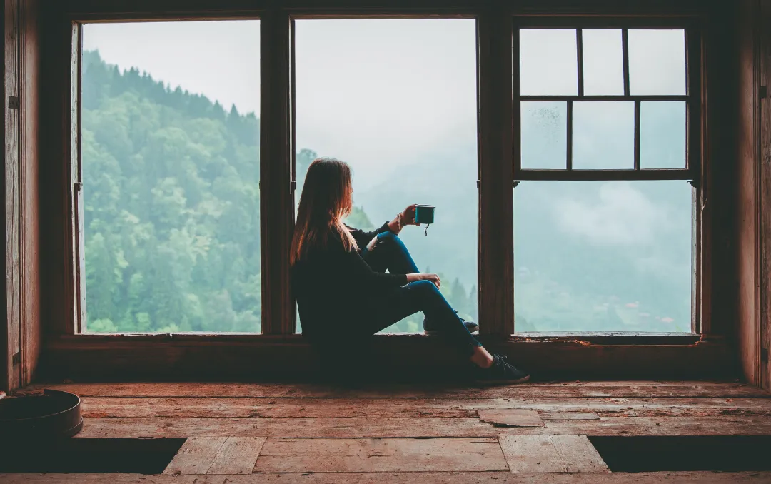 A woman holding a coffee cup gazing out the window into the mountains.