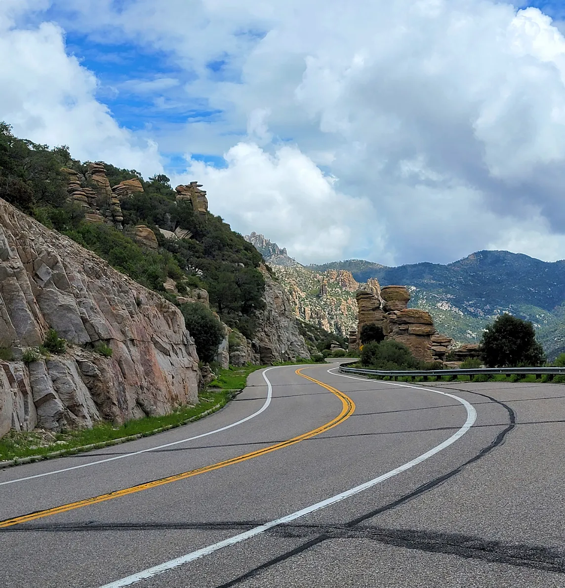 A winding highway going through hoodoo rock formations with green mountains and clouds in the background
