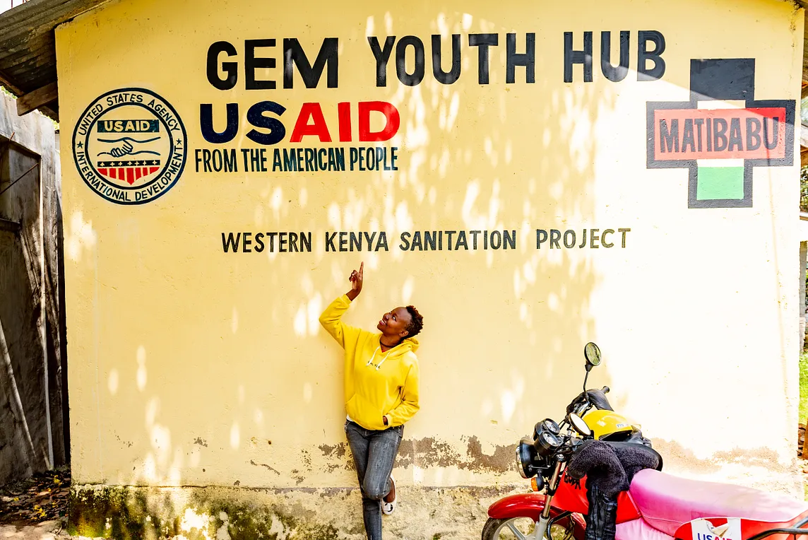 A smiling woman in a yellow sweatshirt stands next to a red motorbike in front of a wall and points to painted words on the wall that say: “Gem Youth Hub USAID. From the American People. Western Kenya Sanitation Project.”