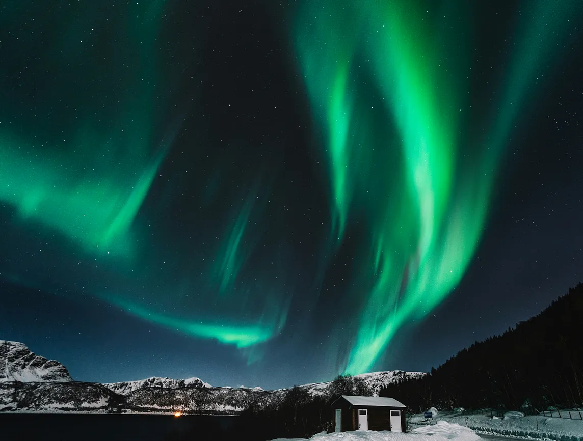 Stunning display of the Northern Lights, or Aurora Borealis, illuminating a clear night sky with vibrant green waves. The bright auroras dance over snow-covered mountains and a small cabin, creating a serene winter landscape in the foreground. Stars dot the dark sky, adding to the ethereal beauty of this natural phenomenon.