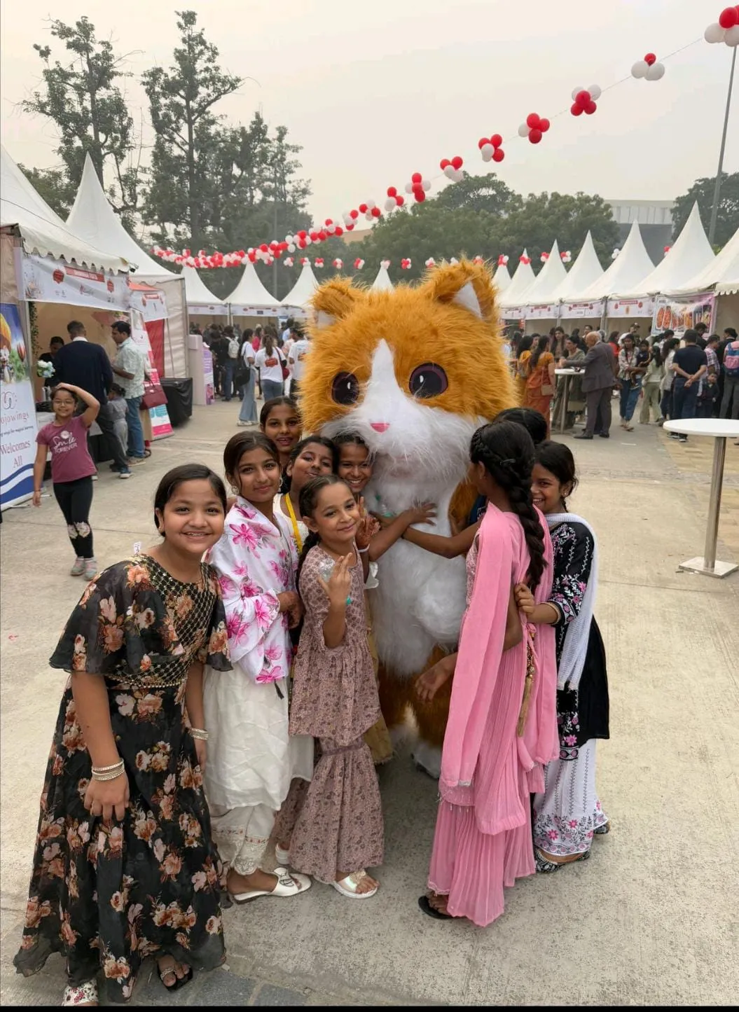 The street students of the NGO at a kids fair. They’re dressed in vibrant clothes and hugging and laughing with a yellow hamster mascot.