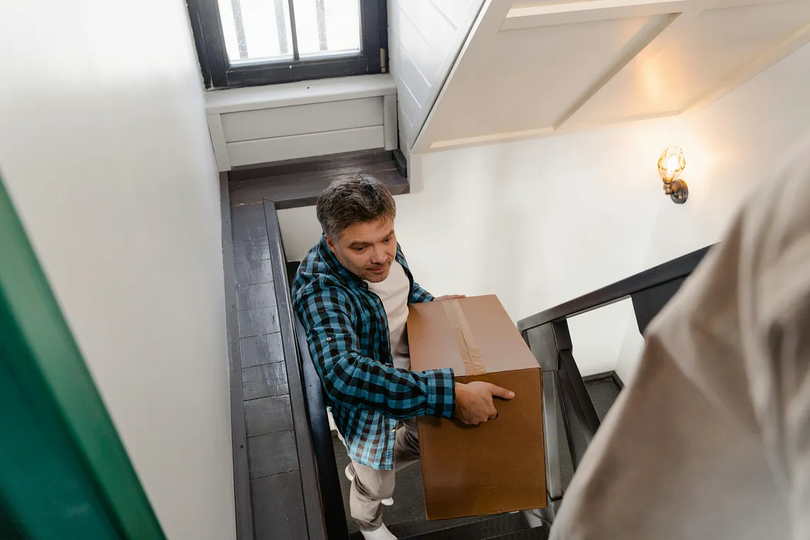 A Man in a checked shirt carrying a large, heavy box up a flight of stairs.