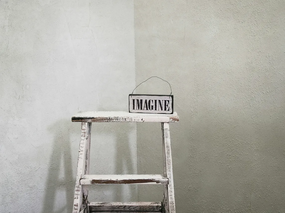 A white painted ladder has a sign on it that reads, “IMAGINE.”