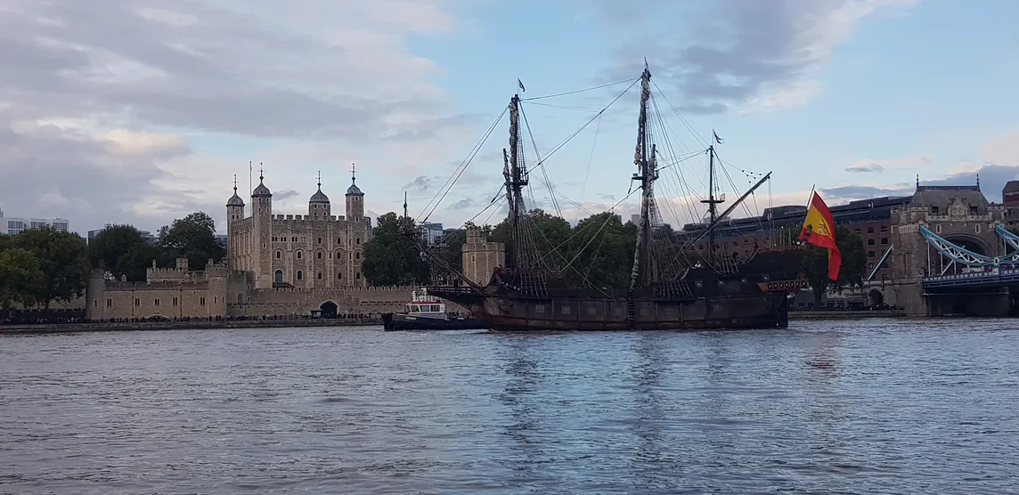 Old tall ship on a river flying a red and yellow flag, passing an ancient large building with several towers