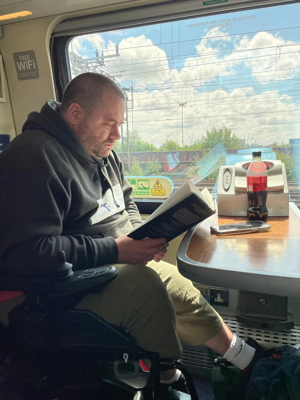 Scott sat reading on a train. On the table in front is a bottle of drink and his phone