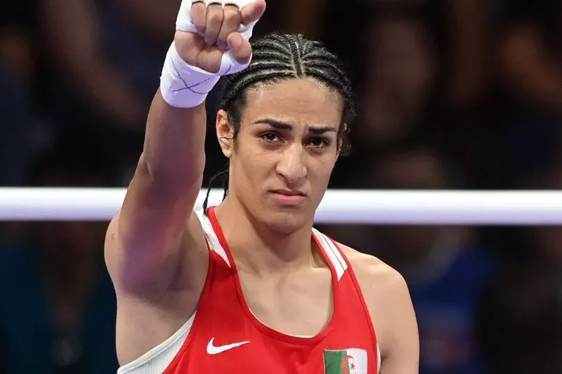 Imane Khelif, a 25-year-old Algerian boxer, is seen in a red boxing uniform with white trim. Her braided hair is tied back, and her hands are wrapped in white tape. She raises her right arm with a clenched fist. The Algerian flag is visible on the left side of her chest while the Nike swoosh logo is seen on the right. She is standing in a boxing ring, with white ropes in the background. Her expression is focused, highlighting her strength and determination.