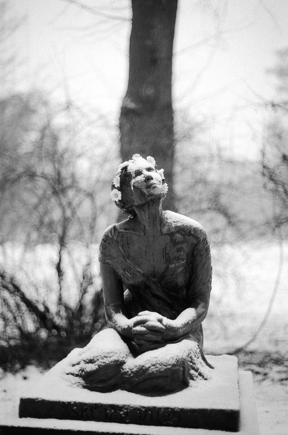 A black-and-white photograph of a snow-covered statue posed in a prayer, looking up to a wintry sky.